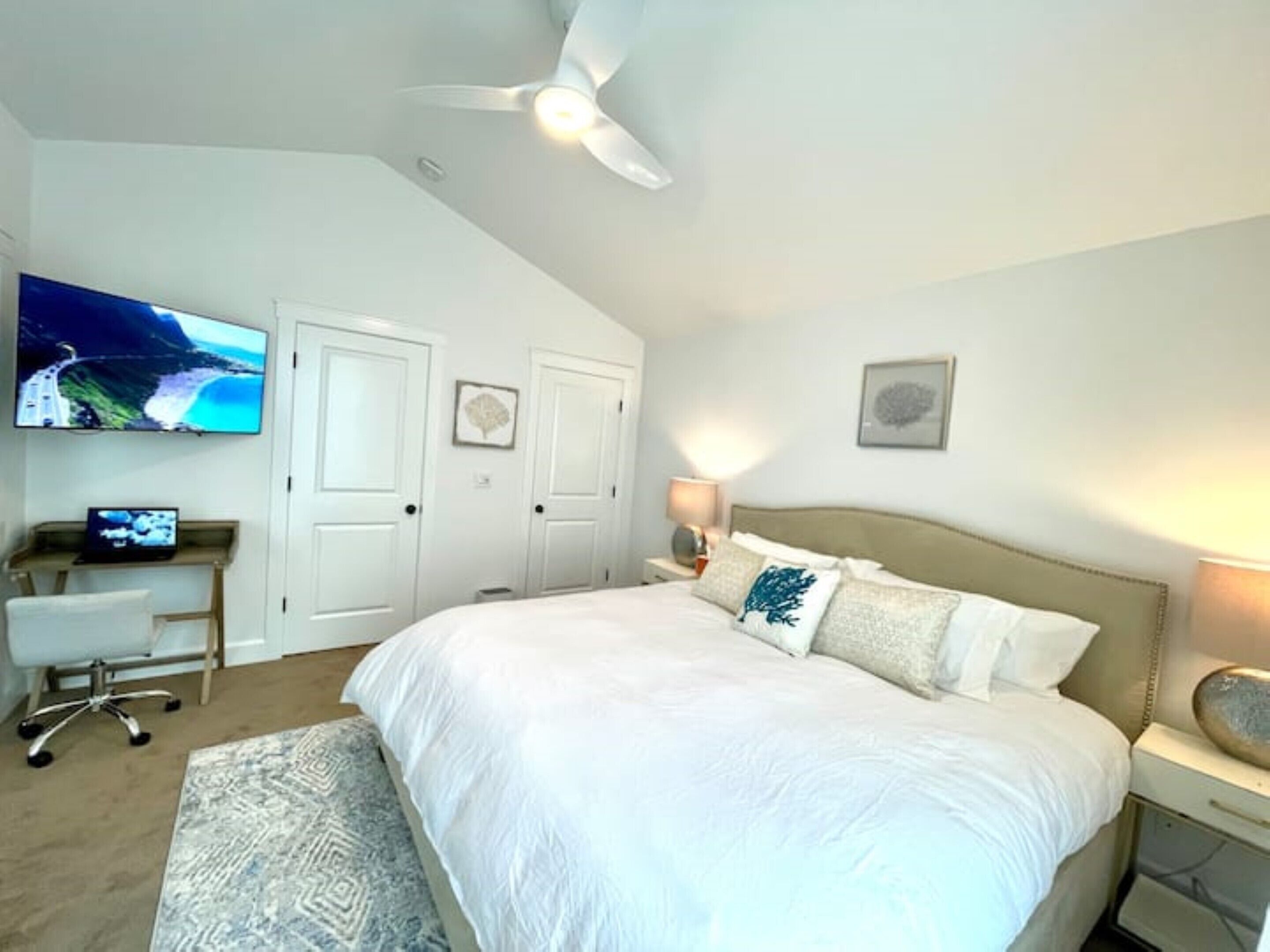 Une chambre avec un grand lit, un bureau avec un ordinateur, un ventilateur de plafond et un téléviseur fixé au mur.