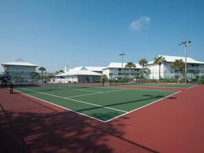 A tennis court with a red and green surface, surrounded by buildings with white roofs and palm trees.