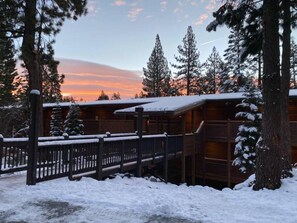 A wooden cabin with a snow-covered deck and a view of the sunset.