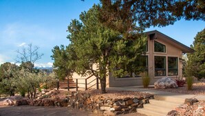 Una casa moderna con un sendero de piedra que conduce a una terraza de madera, rodeada de árboles y un cielo despejado.