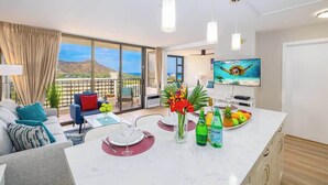 A modern living room with a large TV, a dining area with a fruit bowl, and a balcony with a view. - High-floor Waikiki retreat w/ beach essentials (Honolulu)