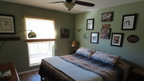 A bedroom with a bed, a ceiling fan, a window with blinds, and various framed pictures on the walls.