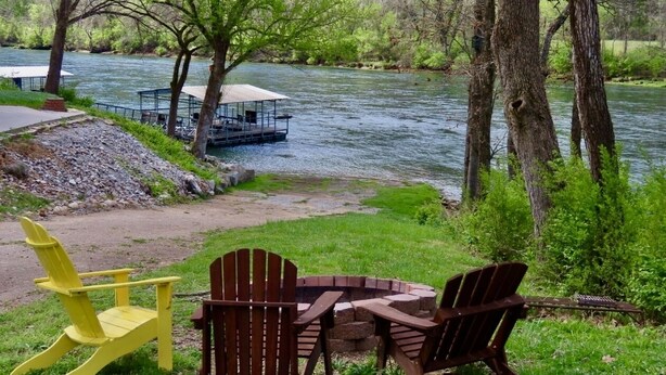 A riverside seating area with Adirondack chairs and a picnic table, overlooking a river and a dock.