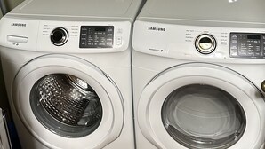 Two Samsung front-loading washing machines in a laundry room with wooden flooring and a light green wall.