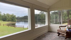 Habitación con amplios ventanales que dan a un lago y una zona verde. Se observa una silla de madera con cojín floral.