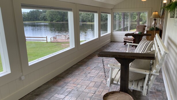Un porche techado con amplios ventanales con vista a un lago, una mesa de madera y un banco.