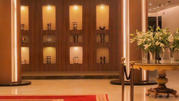 Un hall d’entrée avec un tapis rouge, une table dorée ornée d’un vase de fleurs et des étagères éclairées où sont exposés divers objets.