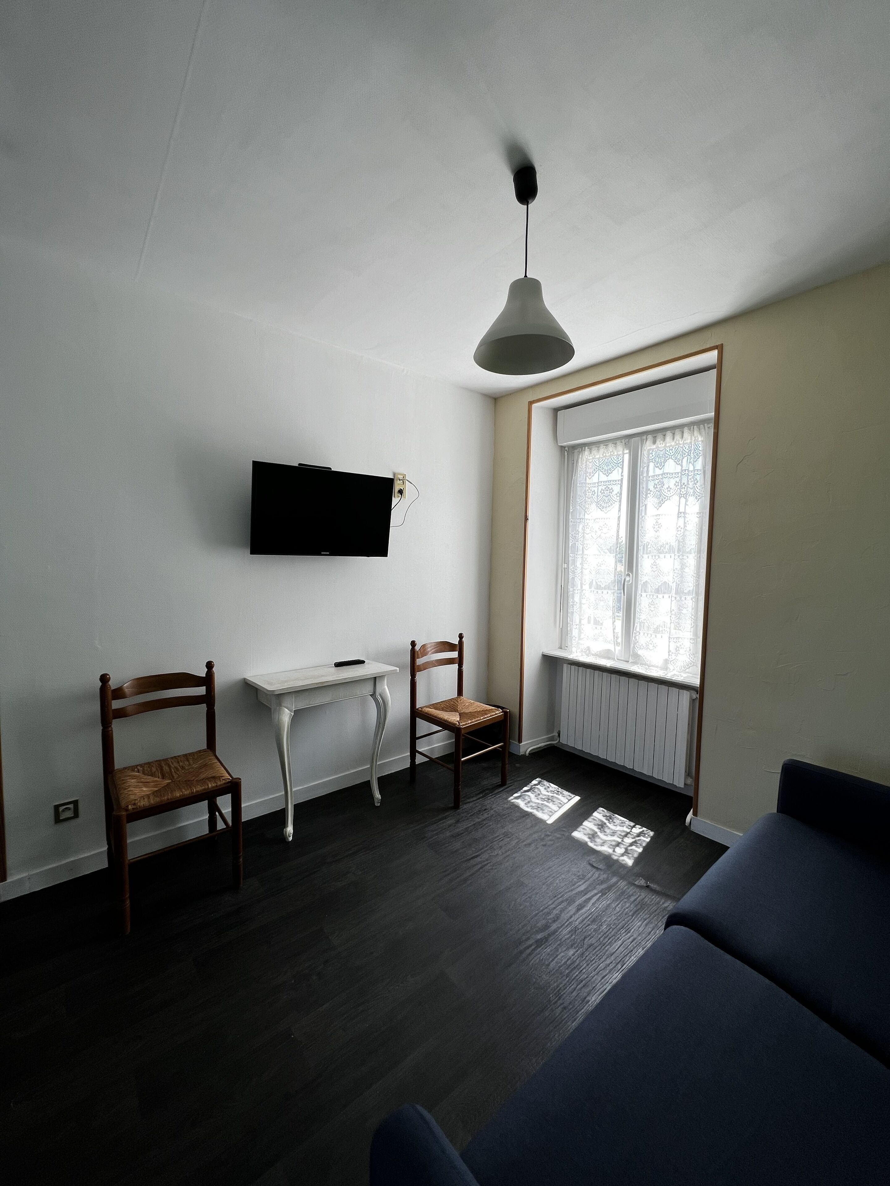 Una sala de estar con piso de madera oscura, una mesa blanca, dos sillas de madera, un sofá azul, un televisor montado y una ventana con cortinas estampadas.