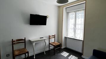 Una sala de estar con piso de madera oscura, una mesa blanca, dos sillas de madera, un sofá azul, un televisor montado y una ventana con cortinas estampadas.
