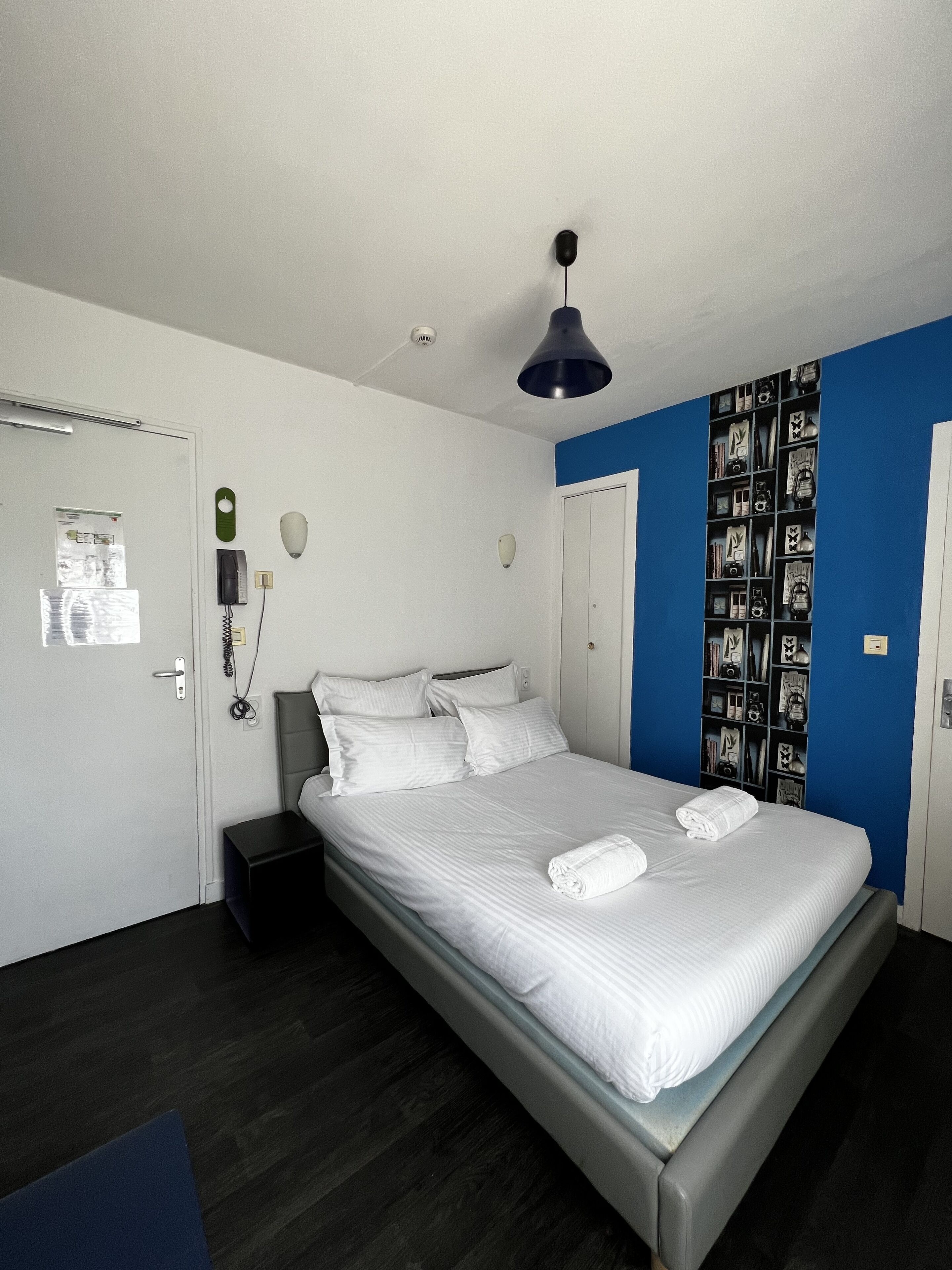 A neatly made bed with white linens and a folded towel, a bedside table, a wall-mounted phone, and a shelf with various items.