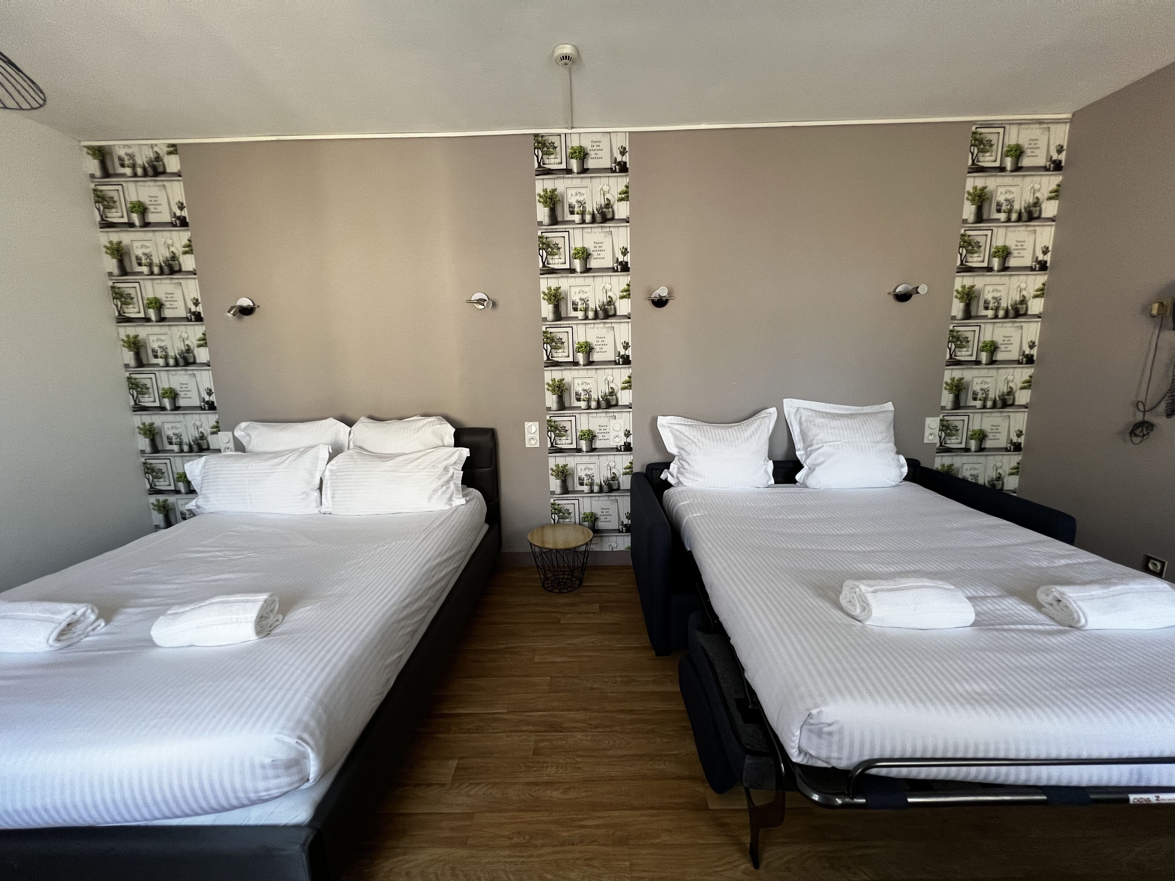 Une chambre double avec deux lits, chacun agrémenté de draps et d’oreillers blancs, une petite table ronde et un mur décoré de tableaux encadrés et de plantes en pot.