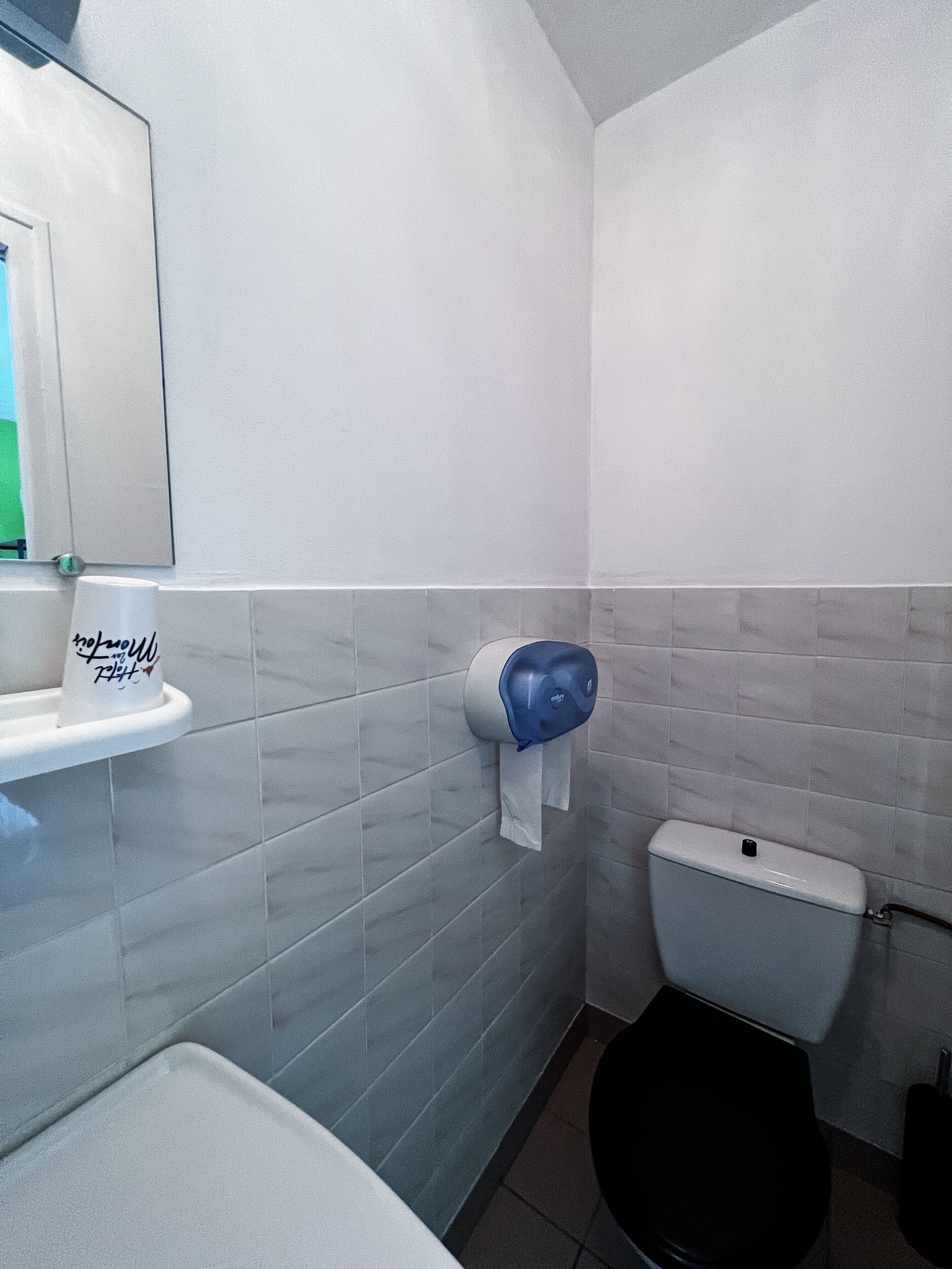 Un baño con azulejos blancos, un inodoro, un lavabo con dispensador de toallas de papel y un espejo.