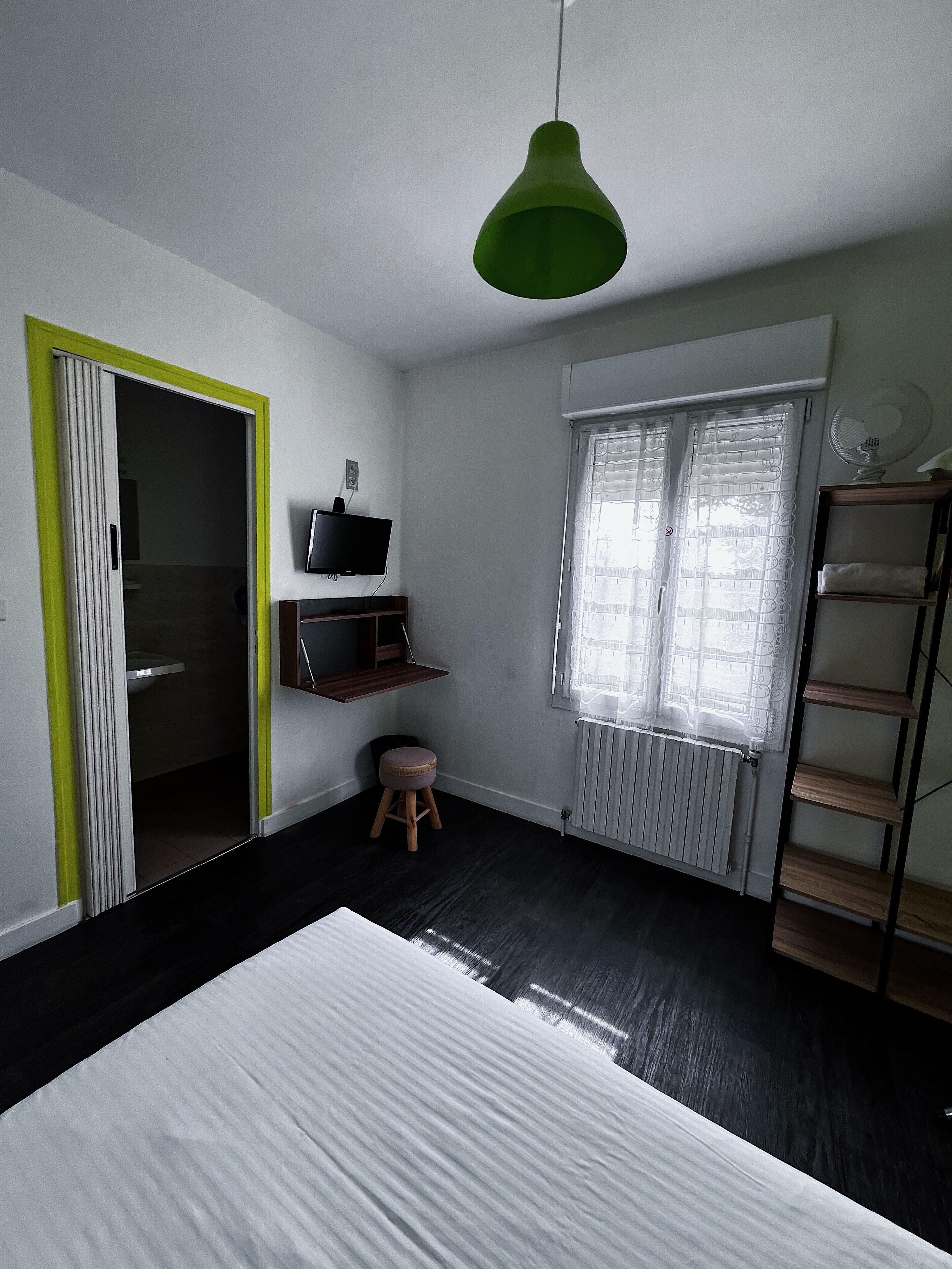 A bedroom with a bed, a green pendant light, a TV, a radiator, a window with blinds, a shelf, and a stool.