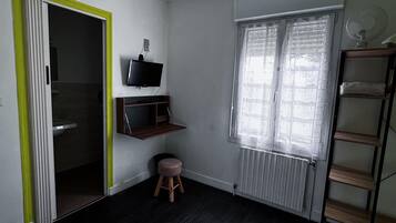 A bedroom with a bed, a green pendant light, a TV, a radiator, a window with blinds, a shelf, and a stool.
