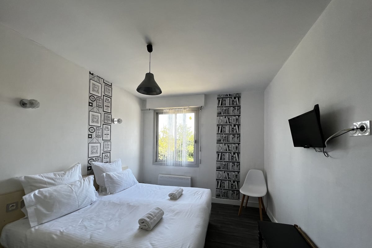 A hotel room with a bed, a desk, a chair, a TV mounted on the wall, and a window with curtains.