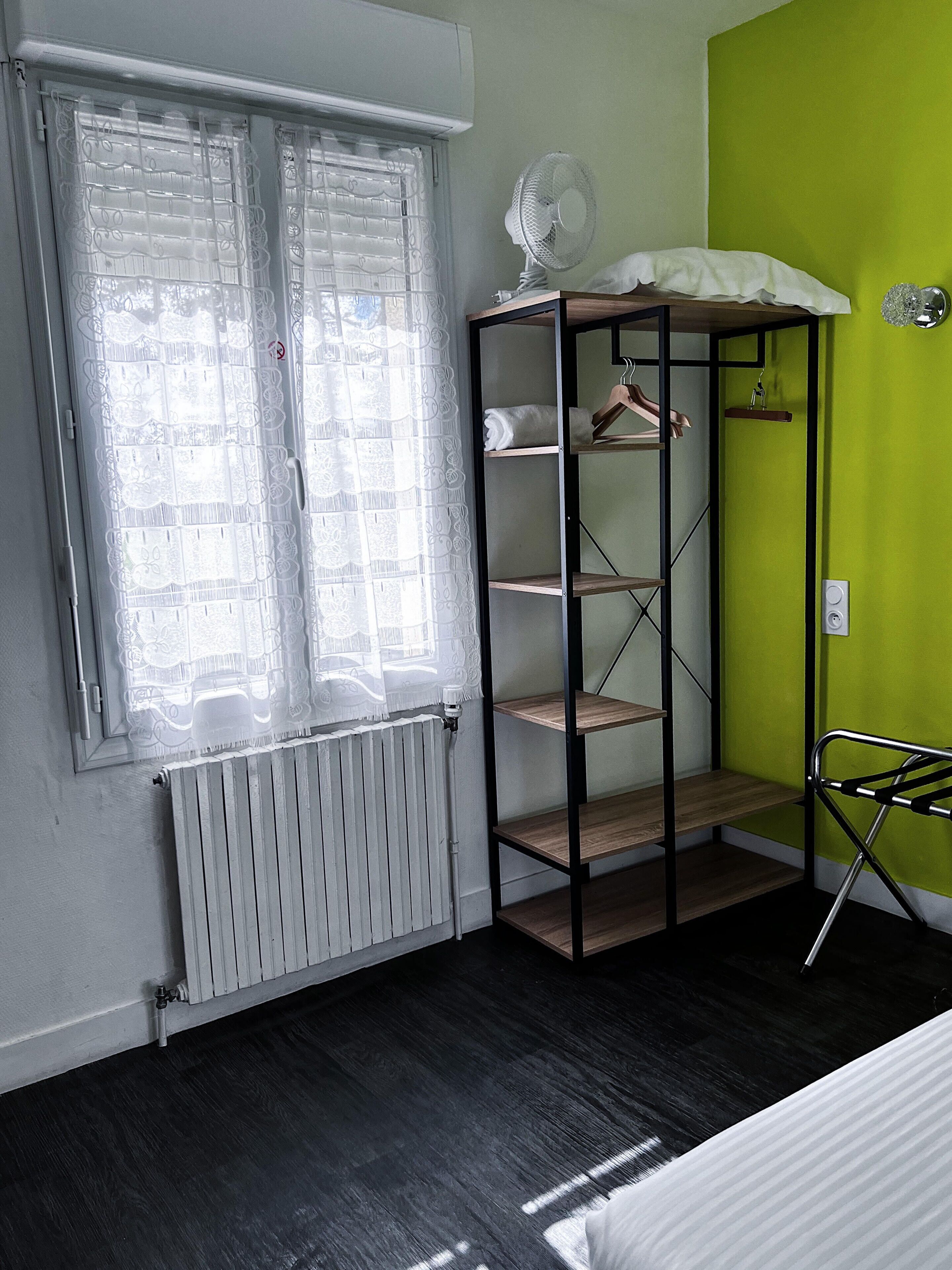 Ein Zimmer mit Heizkörper, einem Vorhangfenster, einer grünen Wand und einem Metallregal mit Kleidung und einem Ventilator.