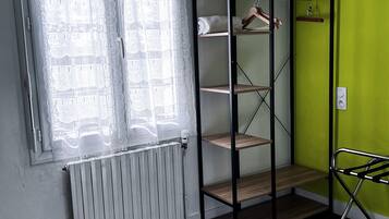 Une pièce avec un radiateur, une fenêtre avec des rideaux, un mur vert et une étagère en métal où sont rangés des vêtements et un ventilateur.