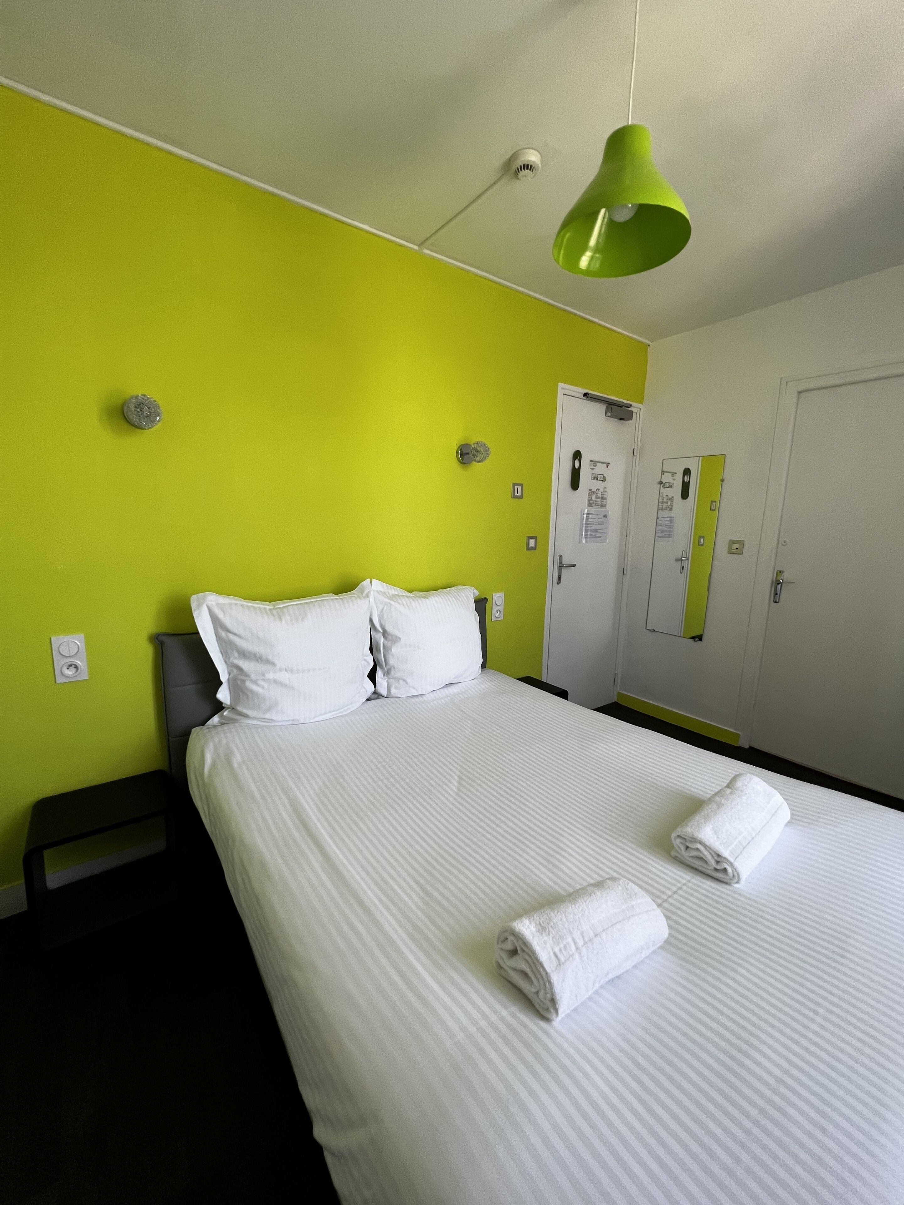 A hotel room with a bed, pillows, a green pendant light, a door, and a towel on the bed.