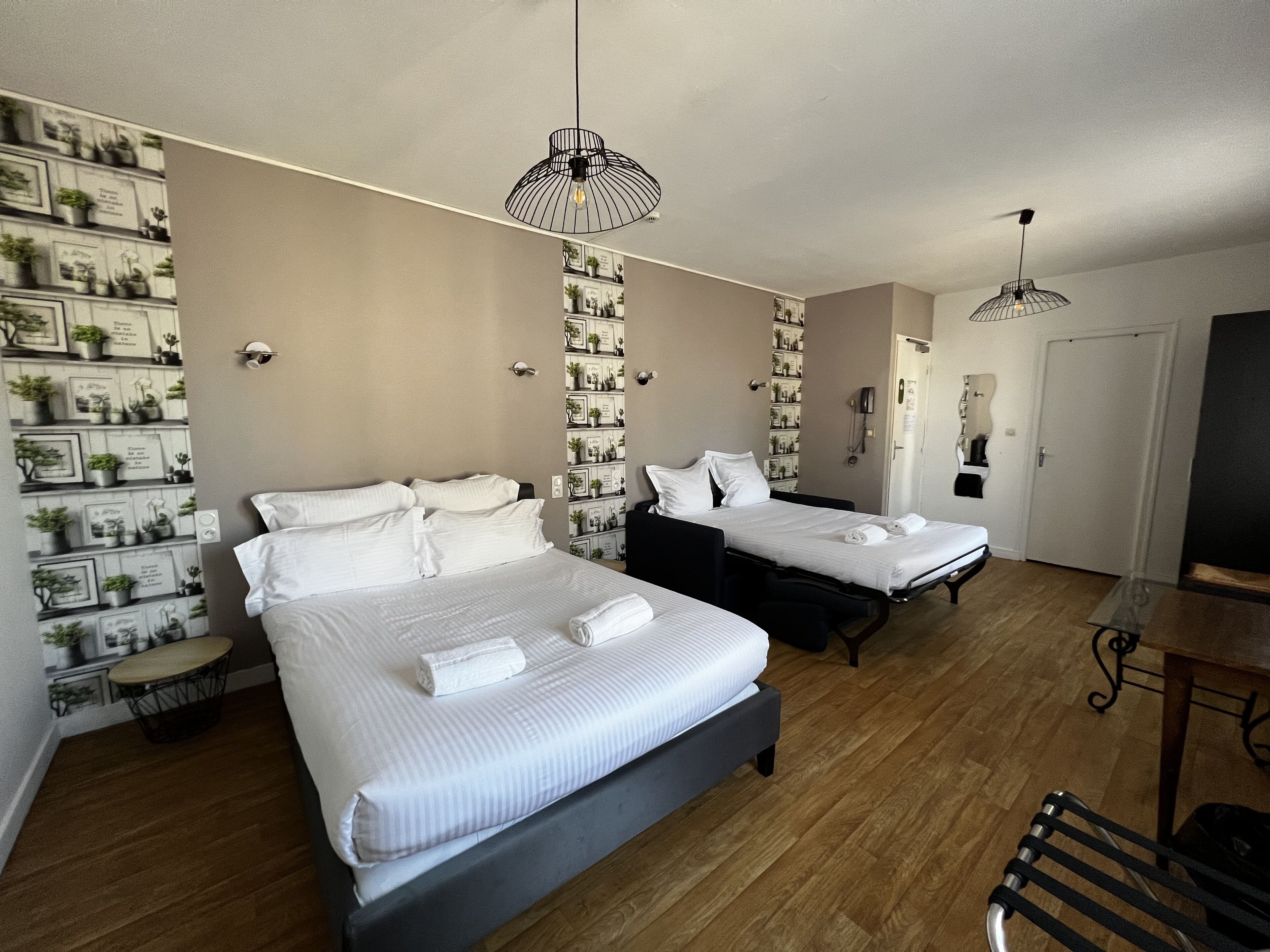 Hotellihuone, jossa on kaksi sänkyä, pieni pöytä, tuoli, seinälle kiinnitetty hylly, jossa on ruukkukasveja, ja kaksi riippuvalaisinta.