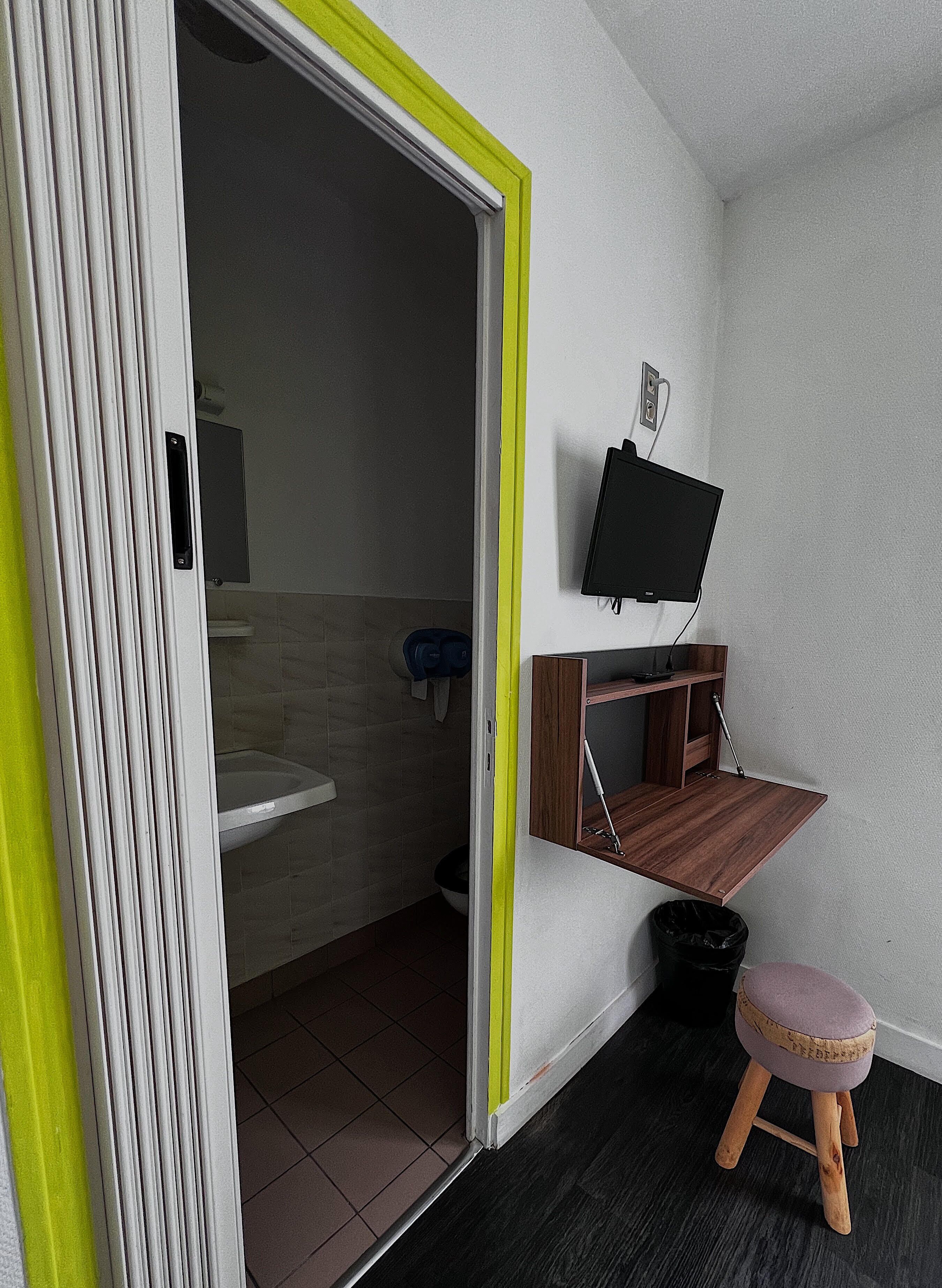 A bathroom with a toilet, sink, and a mirror. There is a television mounted on the wall and a small stool.