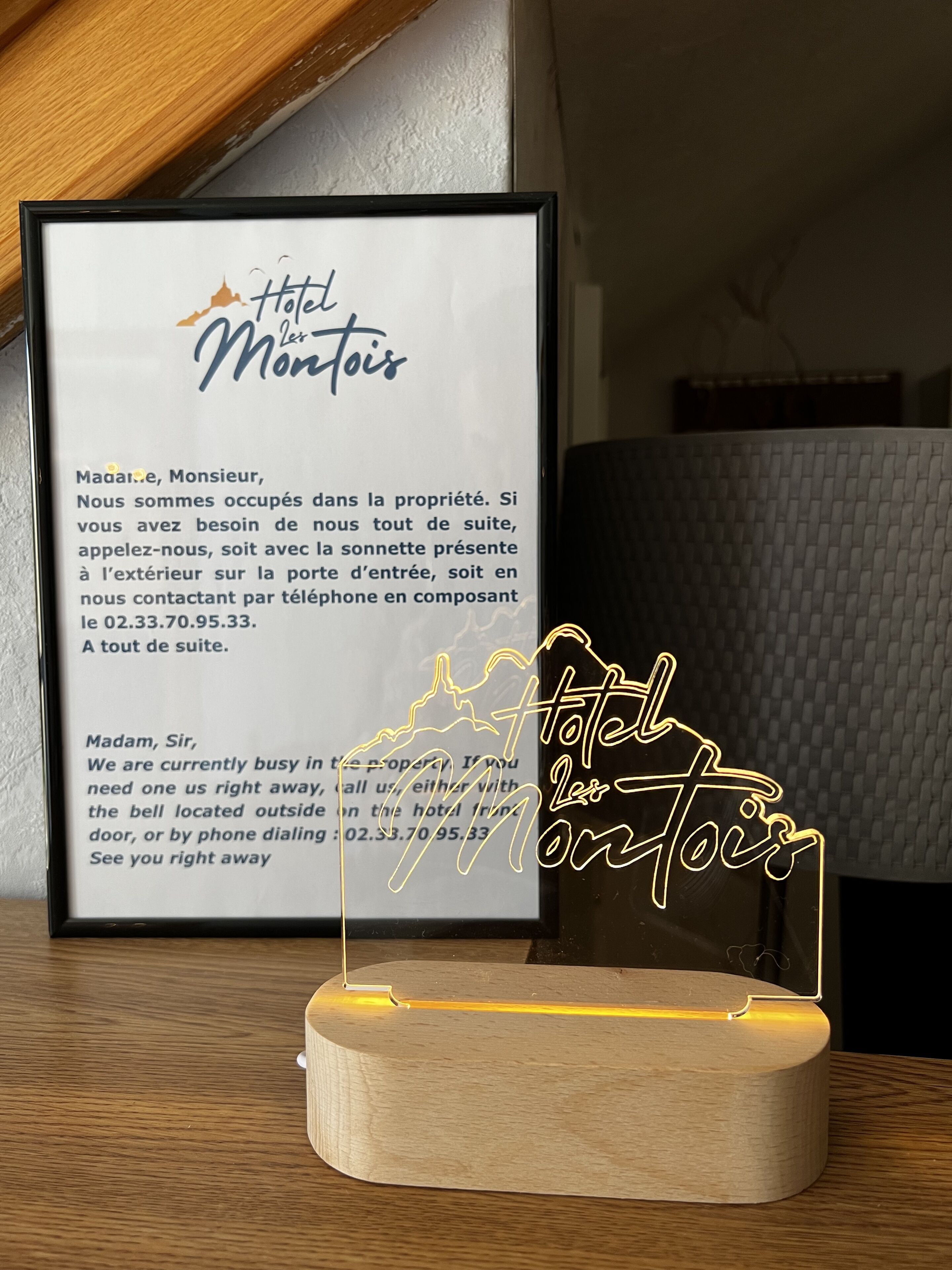 Et rammet skilt for Hotel Montois med kontaktoplysninger og et neontegn foran.