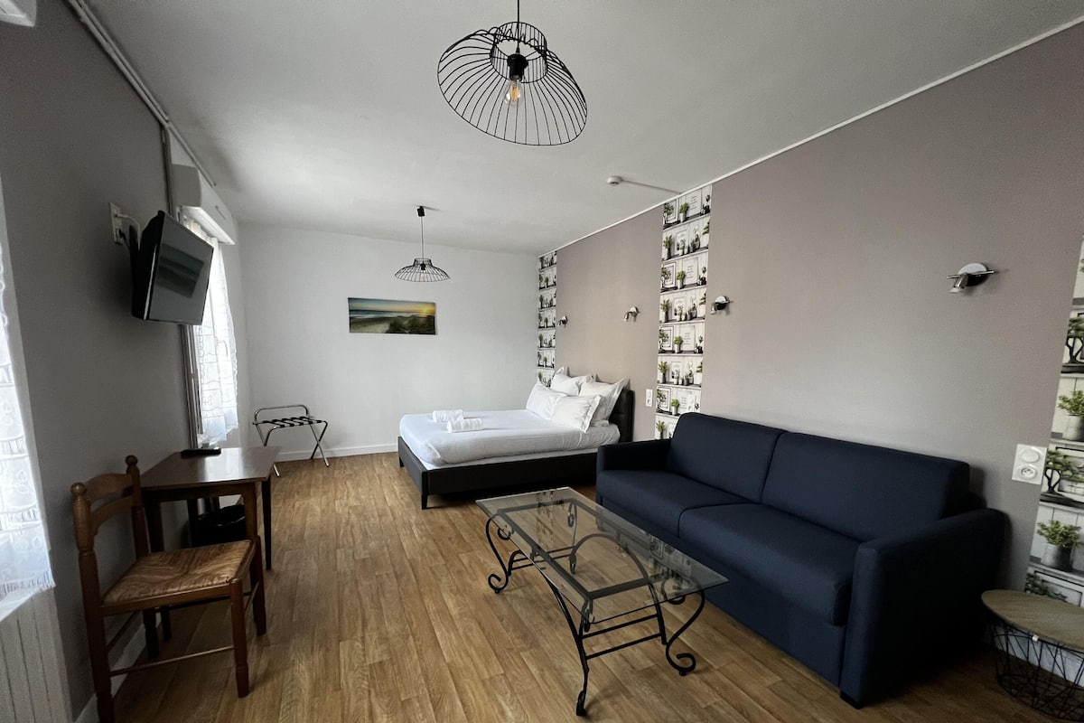 A modern bedroom with a bed, a blue sofa, a wooden dining table, and a TV mounted on the wall.