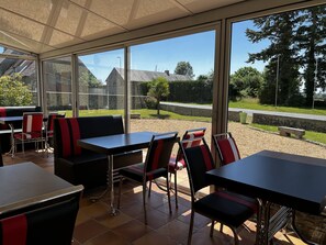 Un espacio de comedor al aire libre con mesas y sillas, ventanas amplias y vistas a una carretera y follaje.