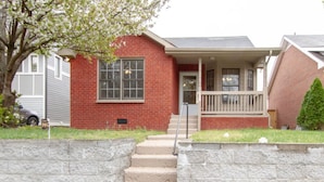 A two-story brick house with a front porch, a visible mailbox, and a small front yard with a tree and a bush. - Bright & Fresh - Large Backyard - Mins to Broadway (Nashville)