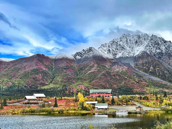 Un albergue con un edificio grande y cabañas más pequeñas, un lago y una cadena montañosa al fondo.