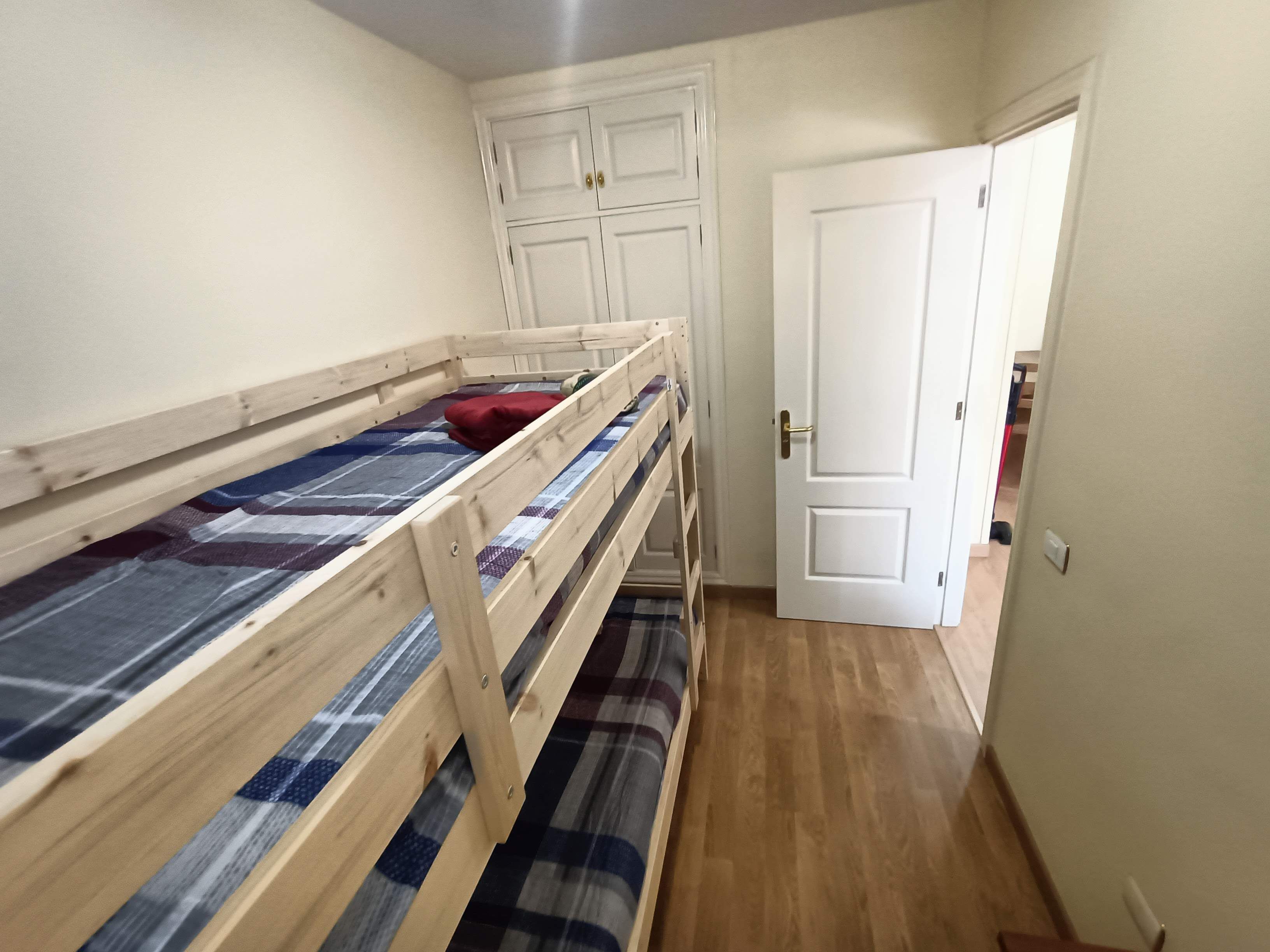 A bedroom with two wooden bunk beds, a white door, and a hallway leading to another room.