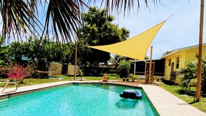 A swimming pool with a yellow shade sail, surrounded by lush greenery and a wooden fence.