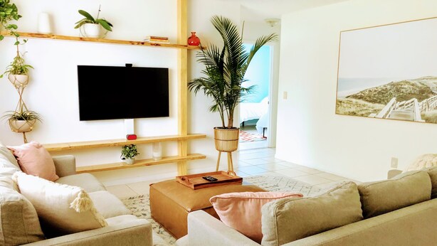 A living room with a flat-screen TV, beige sofas, a coffee table, and various plants.