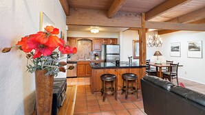 A kitchen with wooden beams, a central island with stools, a refrigerator, and a dining area with chairs and a table. - Fort Marcy 06 | Walk to Plaza | Kiva Fireplace | Full Kitchen | Hot Tub | Pool | Single Level (Santa Fe)
