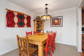 Comedor con mesa de madera y sillas rojas, lámpara colgante, tapiz en la pared y estante con objetos decorativos.