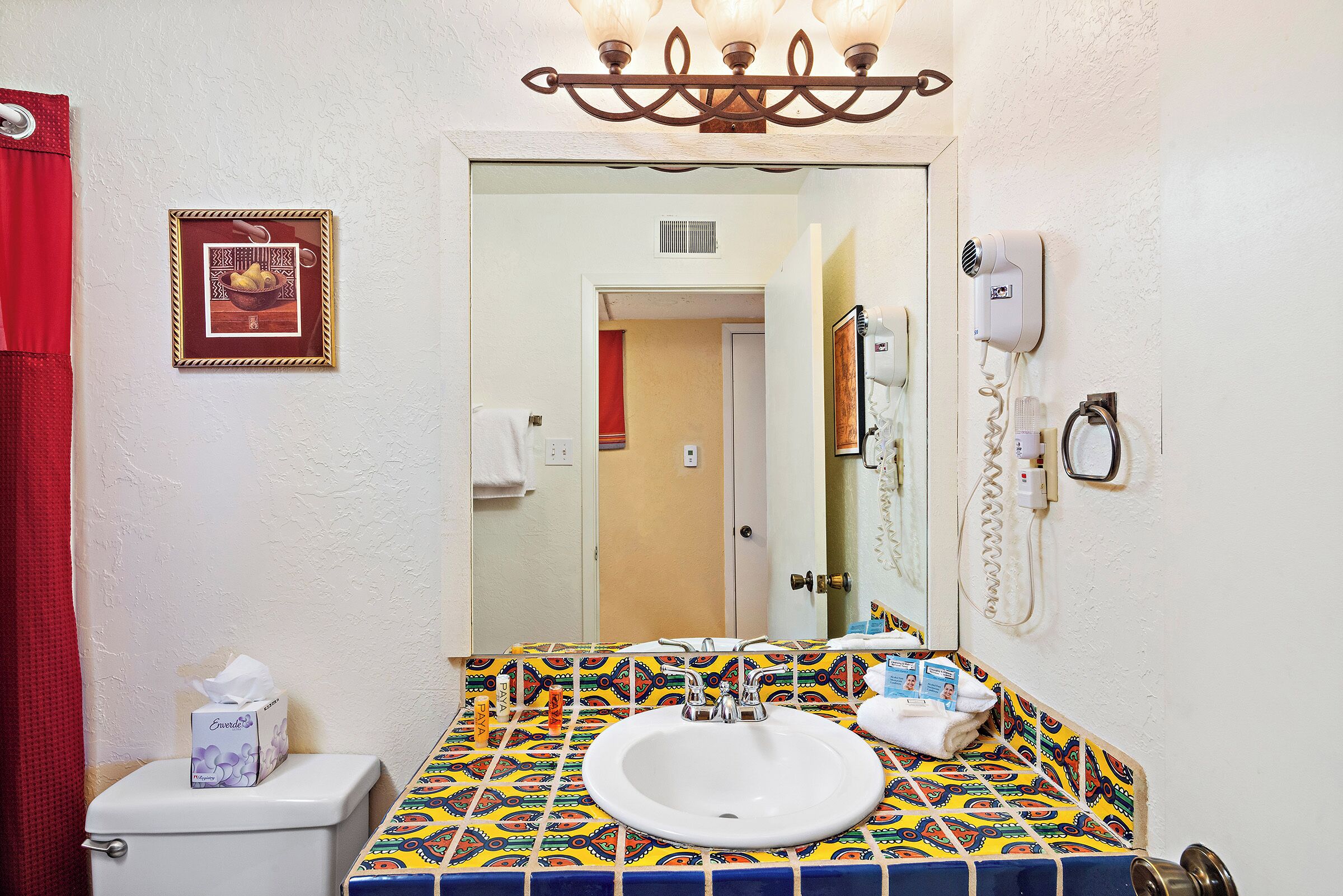 A bathroom with a white sink, colorful tile backsplash, a framed picture on the wall, a toilet, and a mirror with a phone mounted on the wall.