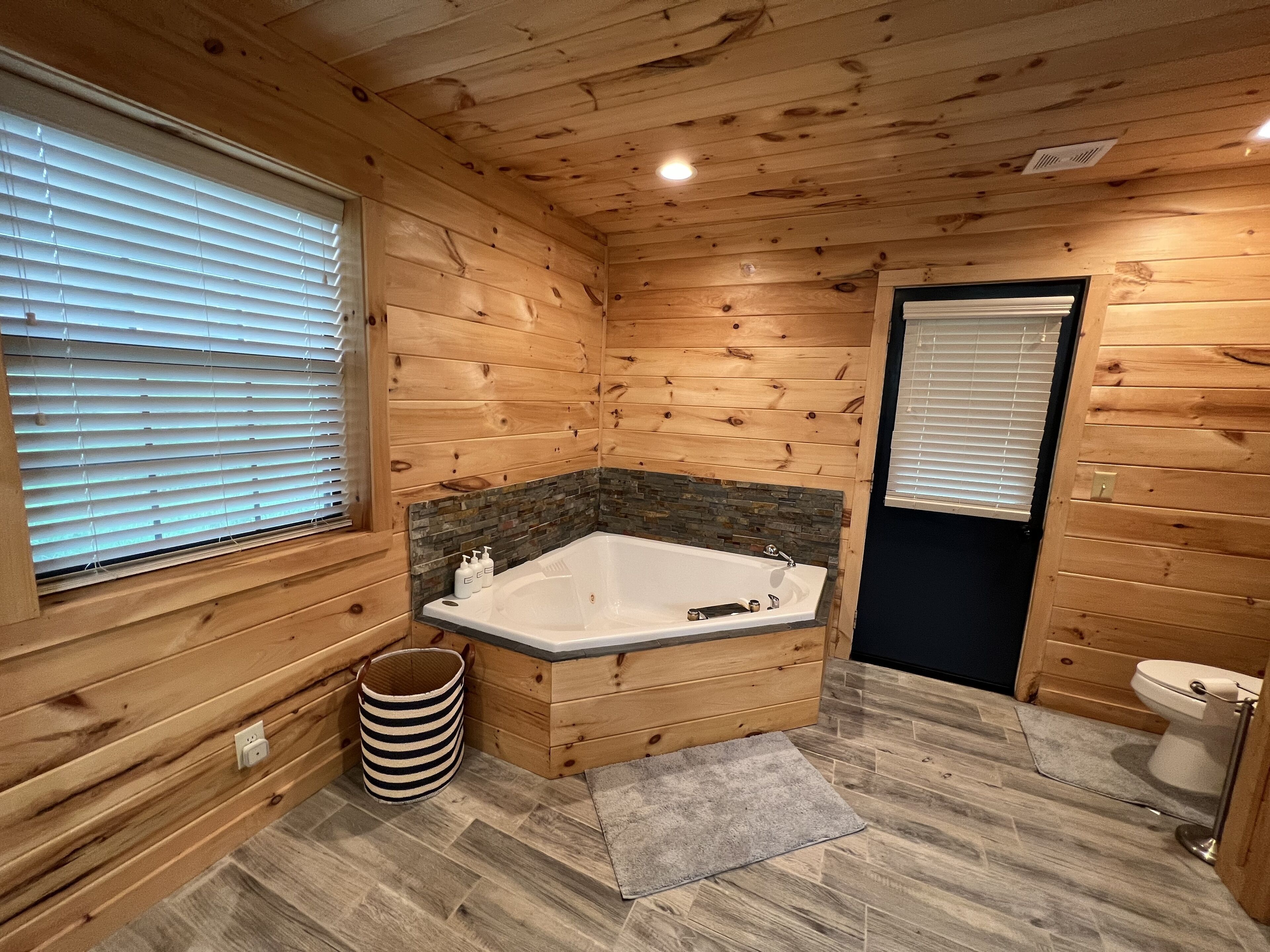 A wooden cabin bathroom with a bathtub, a toilet, and a window with blinds.