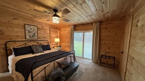 Une chambre à coucher dans une cabane en bois, avec un lit, une table de chevet, une lampe, un ventilateur de plafond, une fenêtre avec des rideaux et une vue sur l’extérieur.