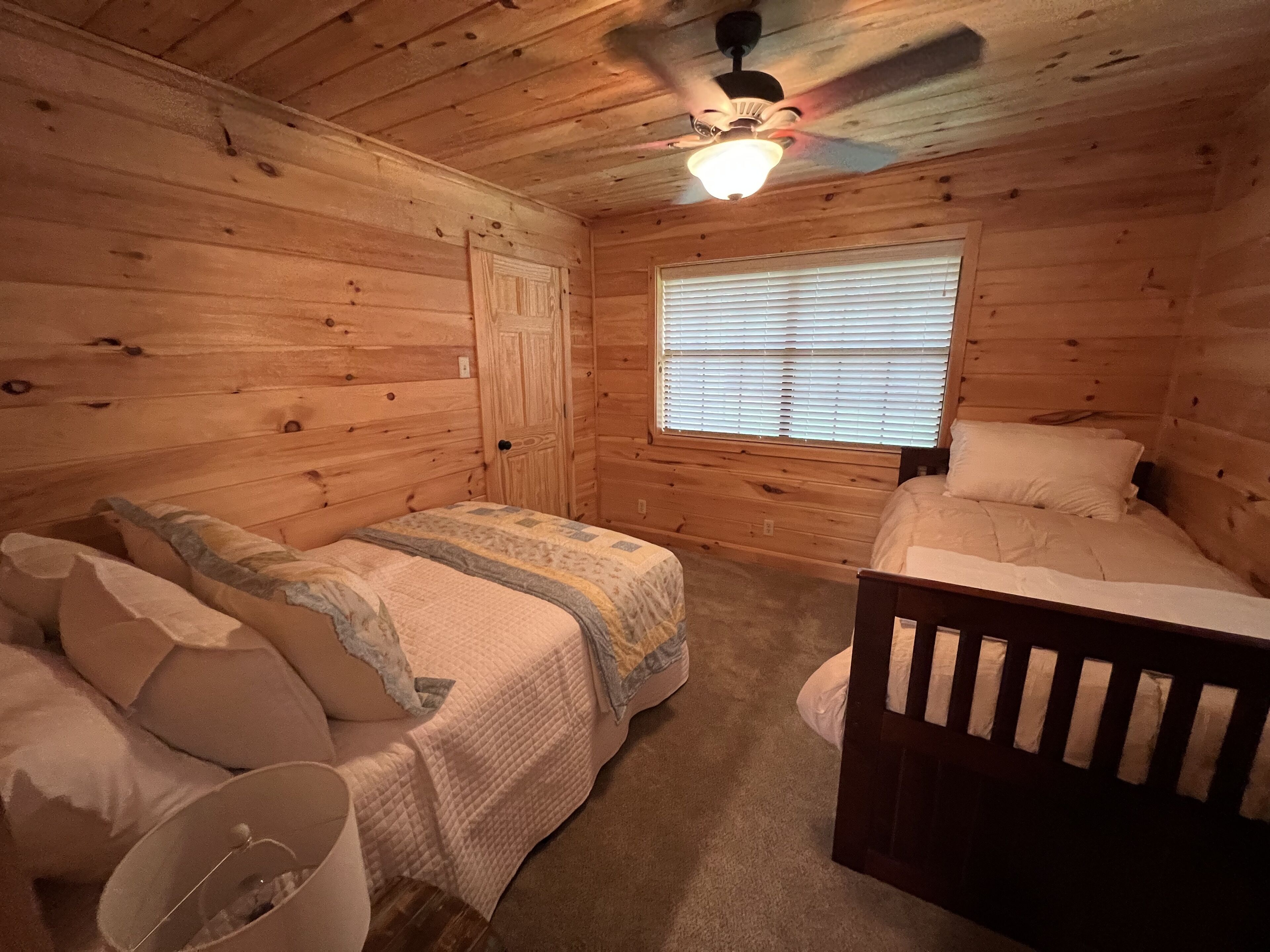 A wooden cabin bedroom with two beds, a ceiling fan, and a window with blinds.