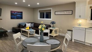 Een moderne woonkamer met een donkerbruine bank, een ronde eettafel met stoelen, een klein wit bijzettafeltje met een bloemenvaas, en een keuken met witte kasten en een vaatwasser.