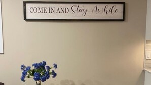 Een kamer met een lijst met een motiverende quote aan de muur, een kleine tafel met een bloemenvaas, een zakdoekendoos en een decoratief object.