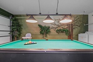 Una mesa de billar con tres lámparas colgantes, una silla blanca y una fila de plantas en macetas frente a una pared verde.