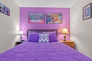 Un dormitorio con colcha morada, dos carteles en la pared y una mesita de noche con lámpara.