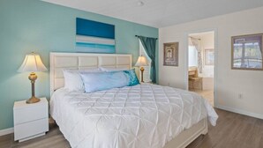 Une chambre avec un lit blanc, deux lampes de chevet, un coussin bleu décoratif et un tableau au mur.