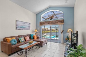 Un amplio salón con un sofá de cuero marrón, cojines coloridos, una mesa de centro de madera y un gran ventanal arqueado con vistas a una piscina.