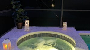 Un bain à remous bordé de carreaux, entouré d’une terrasse en pierre. On peut voir deux lanternes illuminées sur la terrasse, et une plante en pot est visible à l’arrière-plan.
