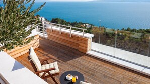 Une terrasse en bois avec une balustrade blanche, une chaise rembourrée et une table sur laquelle se trouvent un verre et un jus d’orange.