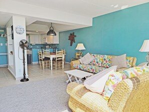 Un salon avec un canapé, une table à manger entourée de chaises, une cuisine en arrière-plan et un ventilateur dans un coin.