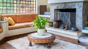 A cozy living room with a stone fireplace, a sofa with cushions, a round wooden coffee table with a plant, and a large window with a view of greenery. - Casa Mantiqueira - Campos do Jordão (Campos do Jordão)