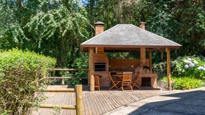 A wooden outdoor barbecue area with a brick chimney, surrounded by greenery and a wooden deck. - Casa Mantiqueira - Campos do Jordão (Campos do Jordão)