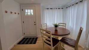 A dining area with a round table, four chairs, a potted plant, a white door, and a window with white curtains.